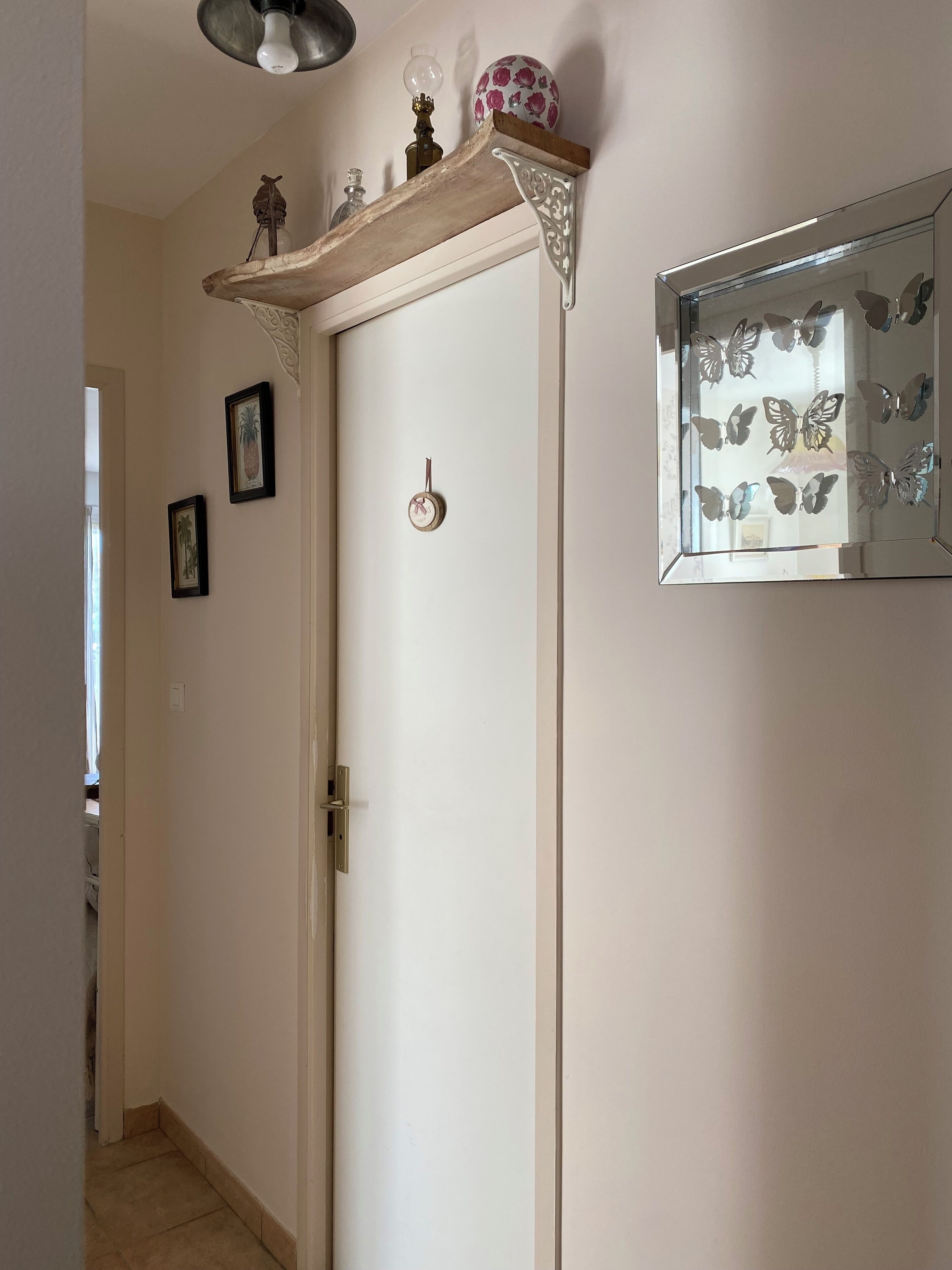 A hallway with a white door, a wooden shelf with decorative items, a wall-mounted mirror with butterfly decals, and framed pictures on the wall.