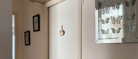 A hallway with a white door, a wooden shelf with decorative items, a wall-mounted mirror with butterfly decals, and framed pictures on the wall.