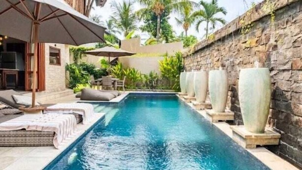 Una piscina con sillas de descanso y sombrillas, rodeada de muros de piedra y palmeras.