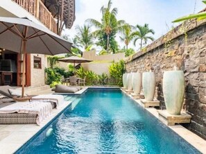 A swimming pool with lounge chairs and umbrellas, surrounded by stone walls and palm trees. - Villa Bambooz Sari (Ubud)