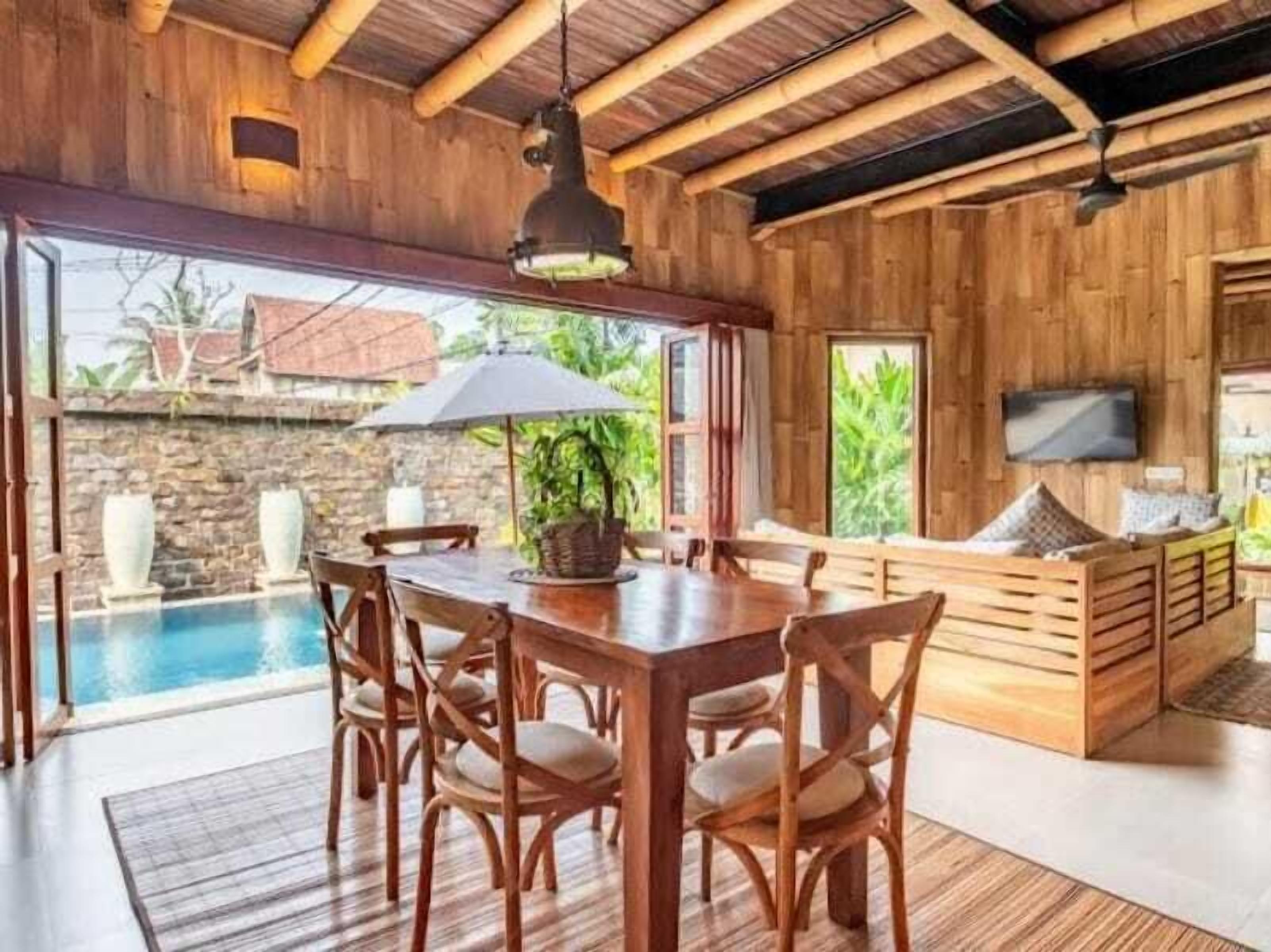 A wooden interior with a dining table, chairs, a sofa, a TV, and a view of a swimming pool.