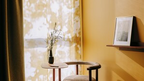 Una habitación con ventana, una silla, una mesita con un florero y un cuadro en la pared.
