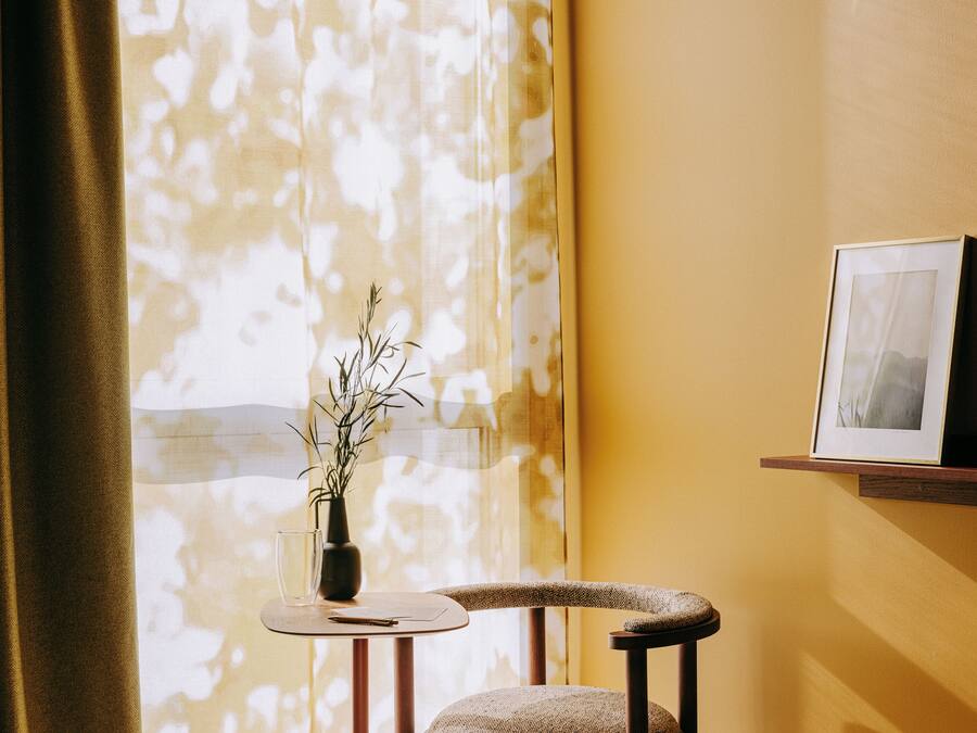 A room with a window, a chair, a small table with a vase, and a framed picture on the wall.