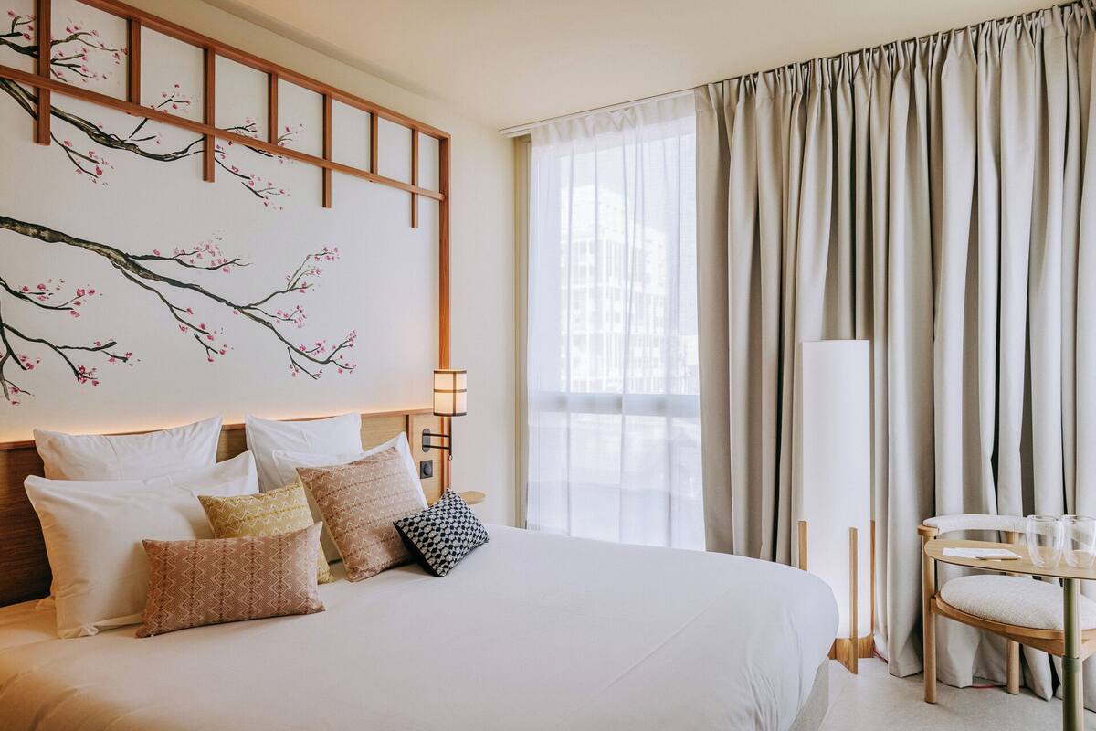 Een hotelkamer met een groot bed, meerdere kussens, een aan de muur bevestigd kunstwerk, een nachtkastje met een lamp, een stoel en een raam met gordijnen.