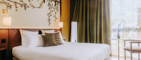 A hotel room with a large bed, a nightstand, a small table with a lamp, a chair, and a view of a balcony with a curtain.