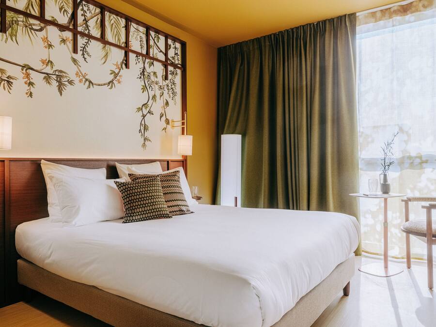 Une chambre d’hôtel avec un grand lit, une table de chevet, une petite table avec une lampe, une chaise et une vue sur un balcon avec un rideau.