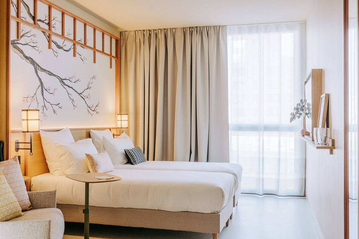 Een hotelkamer met een bed, nachtkastjes, een wandlamp, een groot raam met gordijnen en een decoratief wandobject.