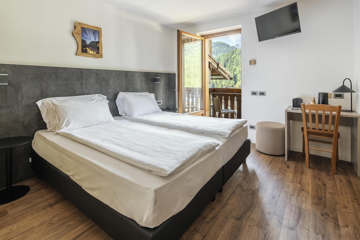 Un dormitorio con cama, mesitas de noche, un escritorio con silla, televisor y ventana con vista a un bosque.