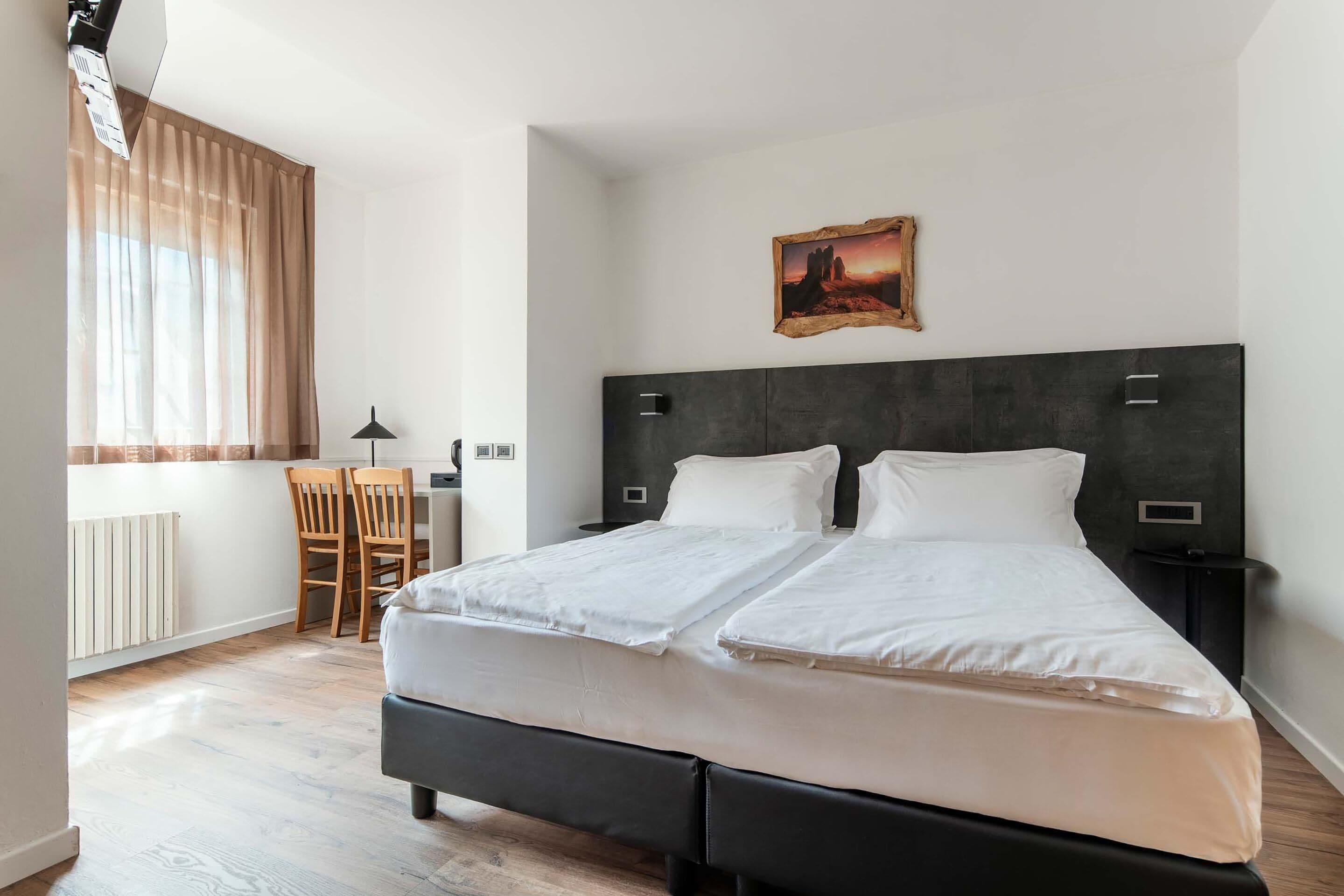 A double bed with a dark headboard, a small desk with a chair, a radiator, and a window with sheer curtains.