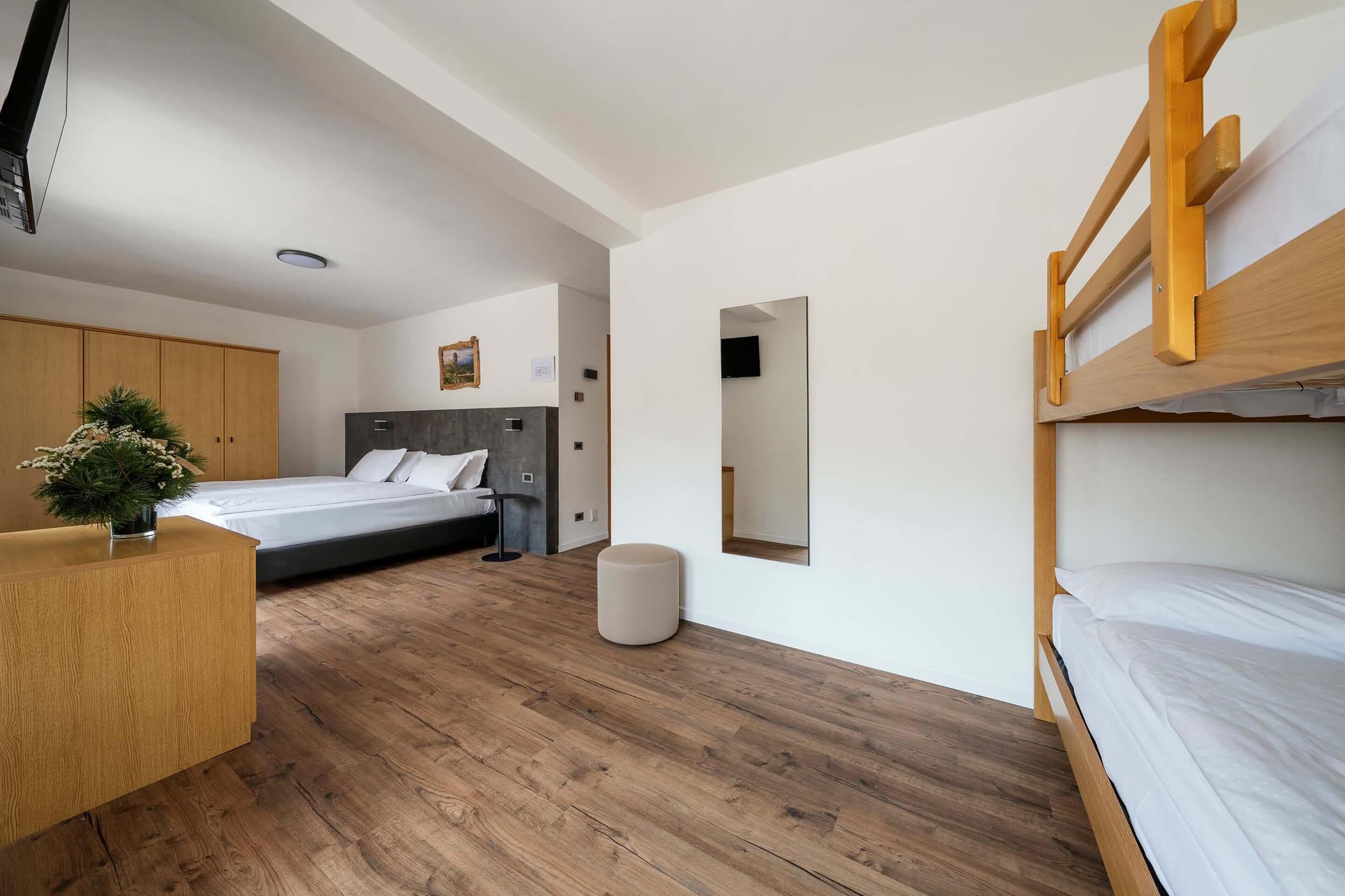 A modern bedroom with a bunk bed, a single bed, a wooden desk, a small potted plant, and a mirror on the wall.