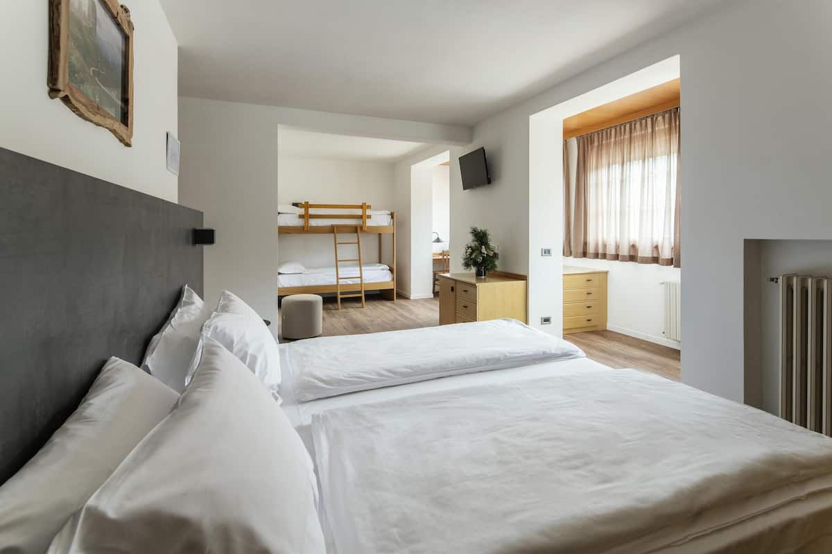 Ein Hotelzimmer mit einem großen Bett, einem kleineren Bett, einem Schreibtisch, einem Stuhl, einem Heizkörper und einem Fernseher an der Wand.