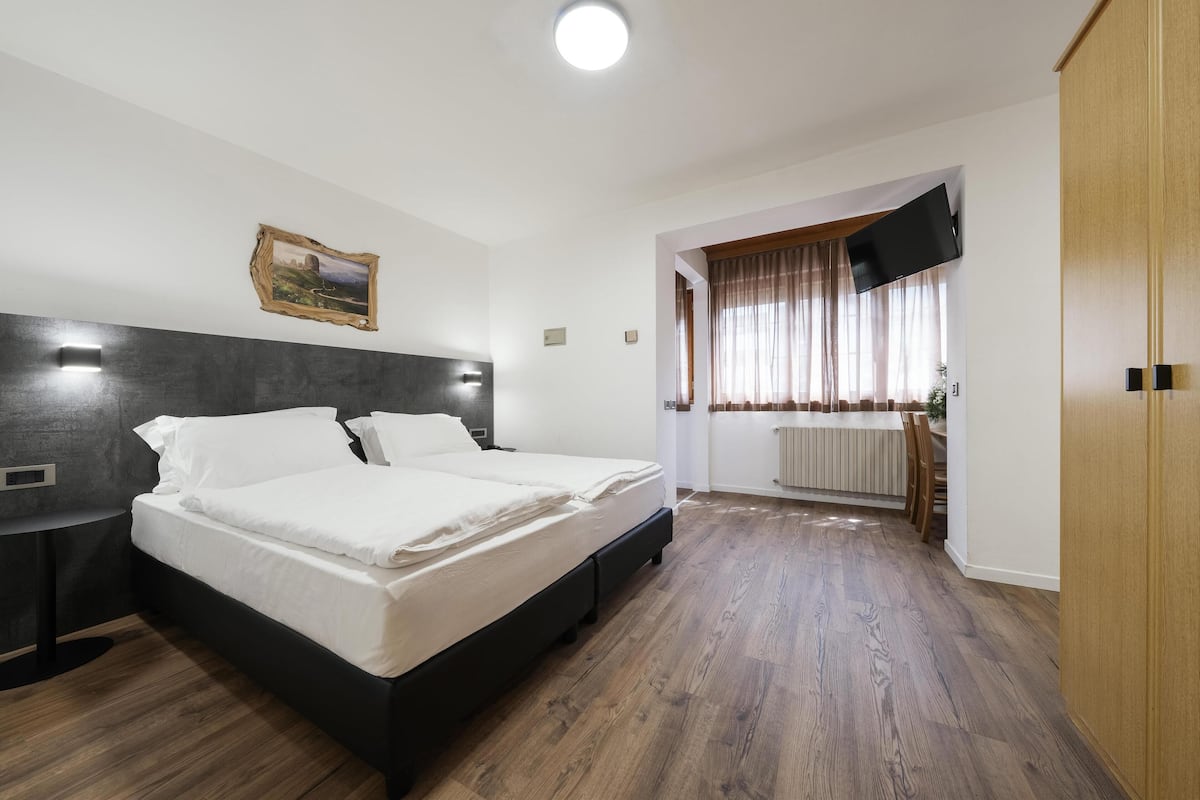 Ein Schlafzimmer mit einem großen Bett, einem Nachttisch, einem an der Wand befestigten Fernseher und einem Fenster mit Vorhängen.