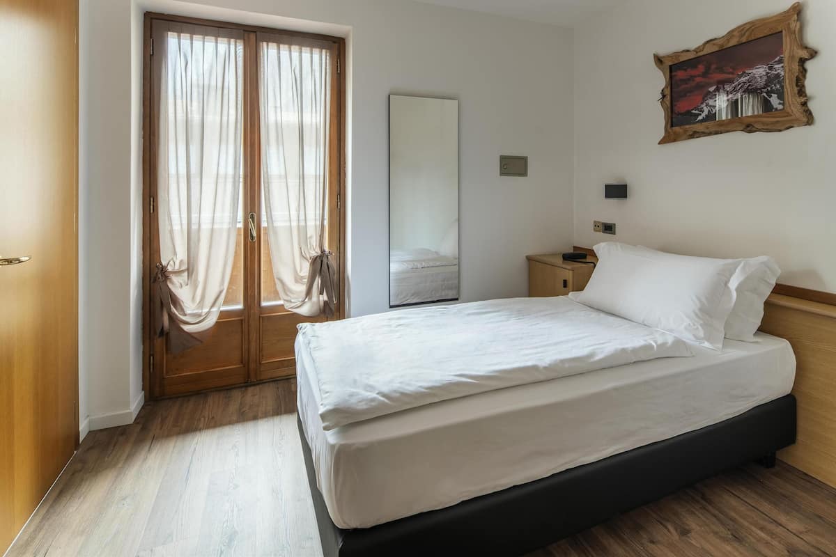 Ein Schlafzimmer mit einem Bett, Holzboden, einer Tür mit Vorhängen, einem Spiegel und einem Gemälde an der Wand.