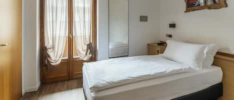 A bedroom with a bed, wooden floor, a door with curtains, a mirror, and a painting on the wall.