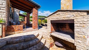 Uma cozinha ao ar livre construída em pedra, com chaminé, uma área de estar coberta com mesa e cadeiras, e uma escadaria de pedra que a conduz.