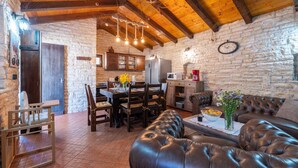 Um interior rústico com parede de pedra, vigas de madeira no teto, área de jantar com mesa e cadeiras, sofá de couro e cozinha equipada.