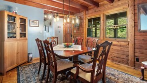 A wooden dining room with a large table, six chairs, a chandelier, and a window with a view of greenery.