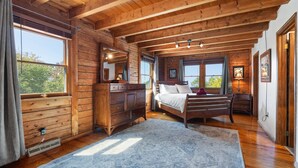 A wooden cabin bedroom with a large bed, a dresser, and a window overlooking greenery.