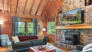 A log cabin interior with a stone fireplace, wooden walls, a large TV, and a cozy seating area.