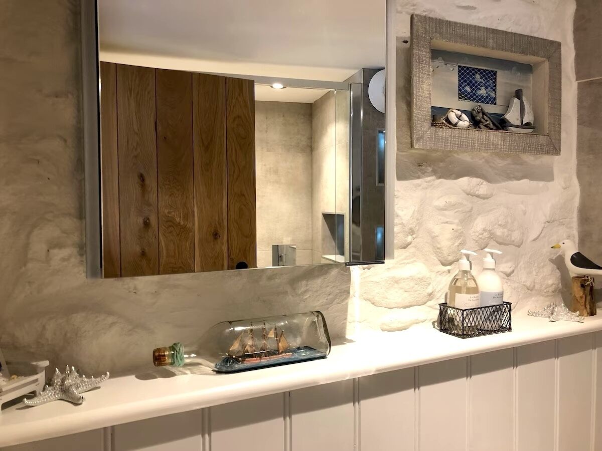 A bathroom with a wooden vanity, a large mirror, a small shelf with decorative items, and a glass jar with a ship model.