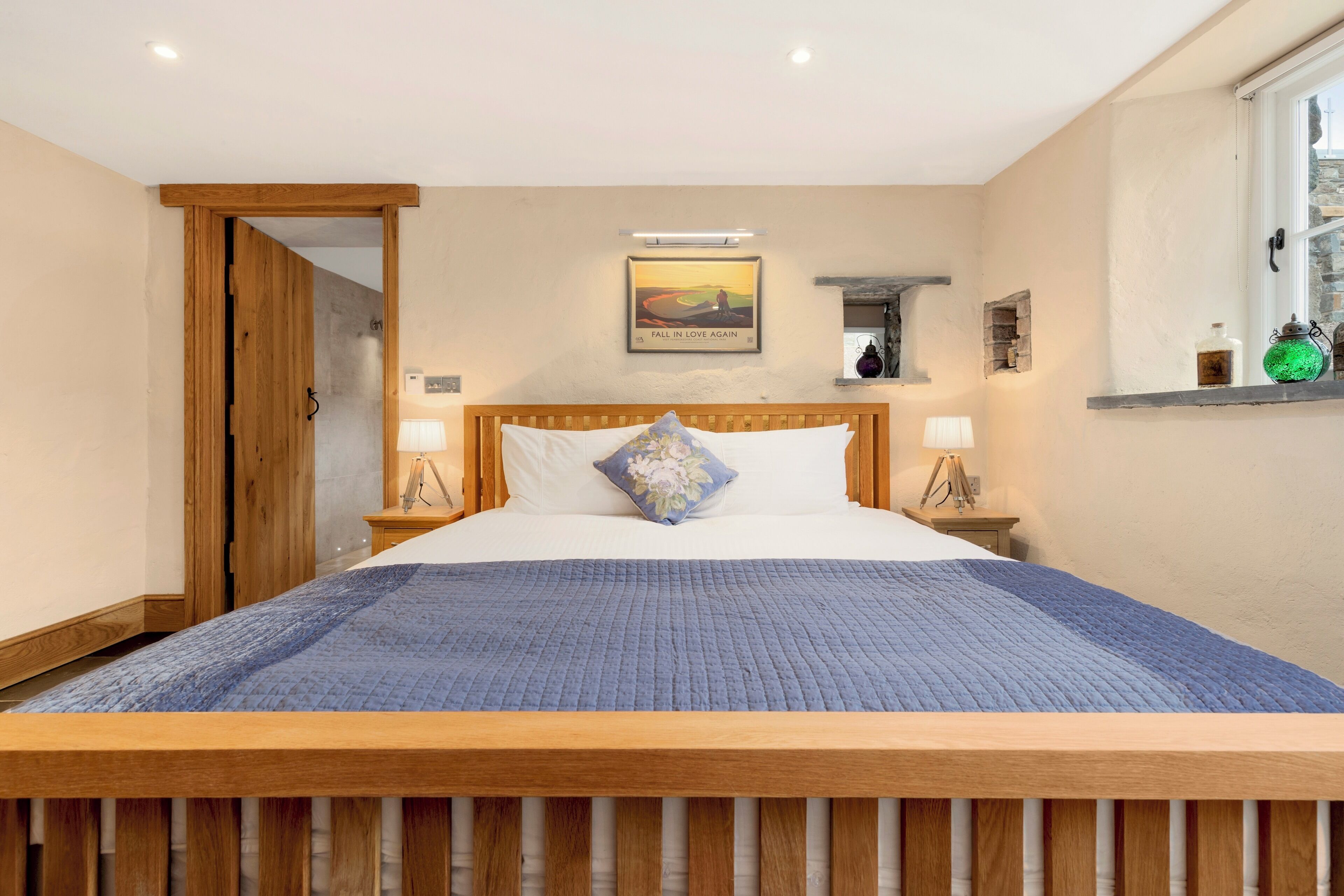 Quarto com cama de madeira, colcha azul, pintura na parede e janela com vista.