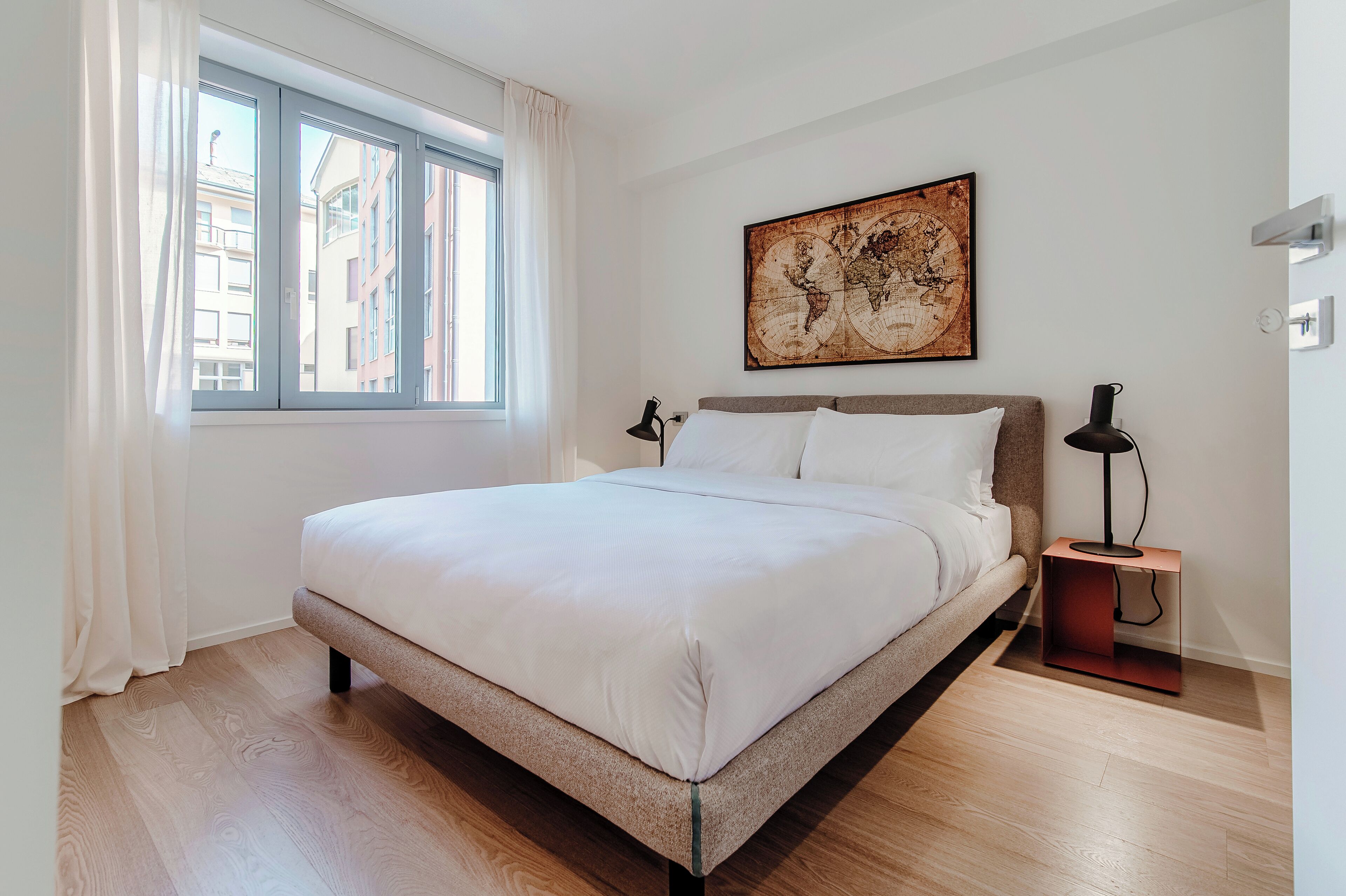 A modern bedroom with a large bed, a world map artwork, a bedside lamp, and a window with curtains.