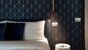 Quarto de hotel com parede de padrão escuro, cama com roupa de cama branca e azul escura, mesa de cabeceira com abajur e tomada na parede.