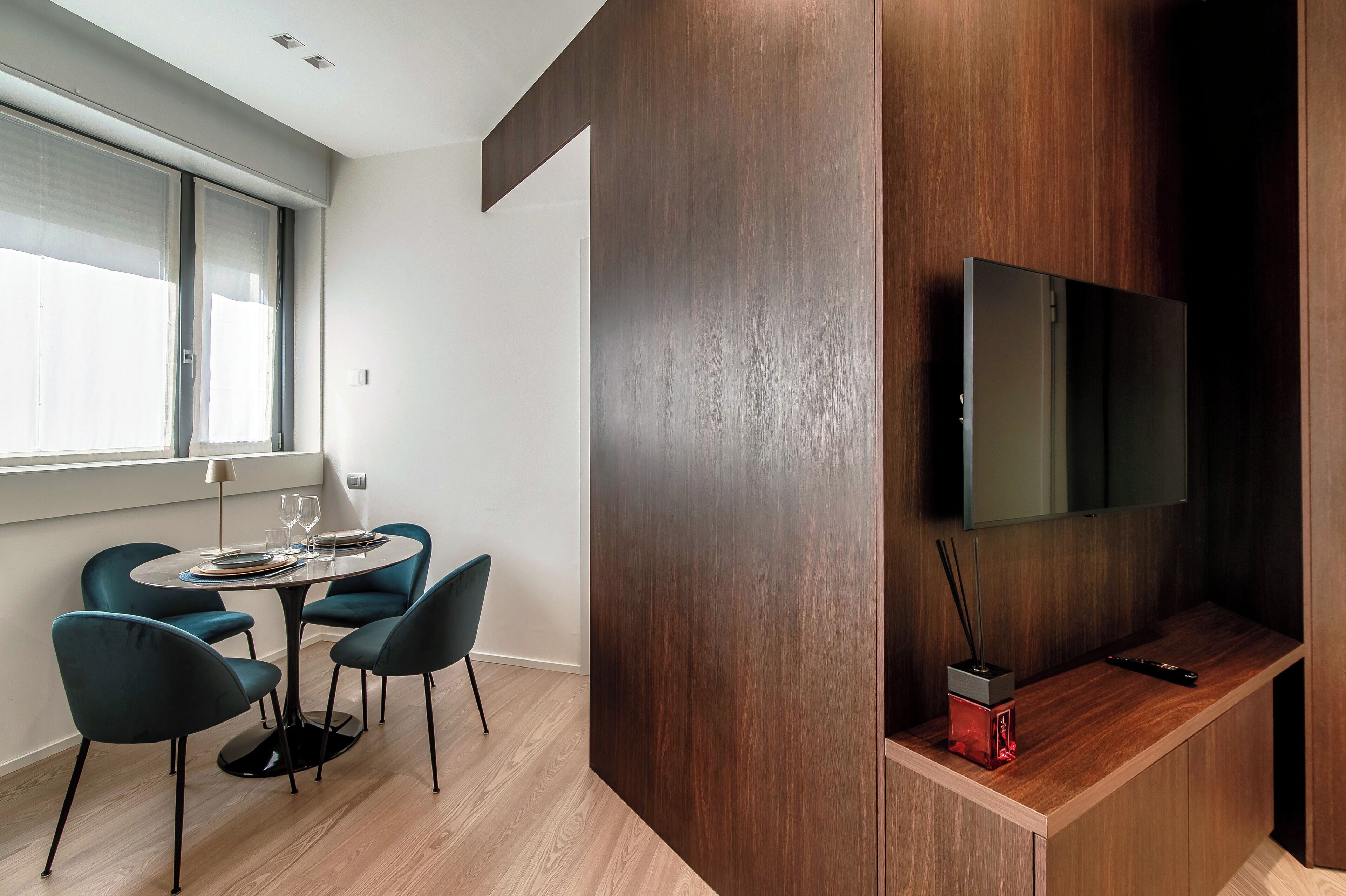 Un comedor moderno con una mesa redonda, sillas color turquesa y una pared de madera con un televisor montado.