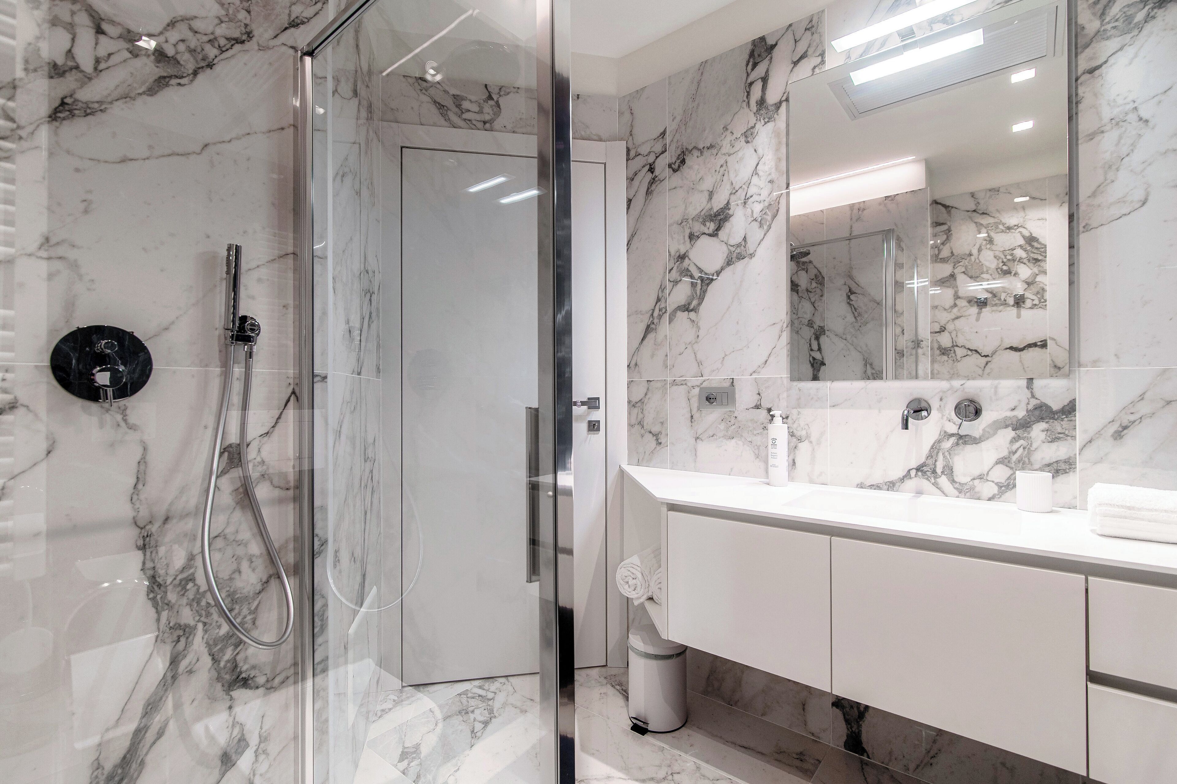 A modern bathroom with a glass-enclosed shower, marble walls, and a large mirror above the sink.