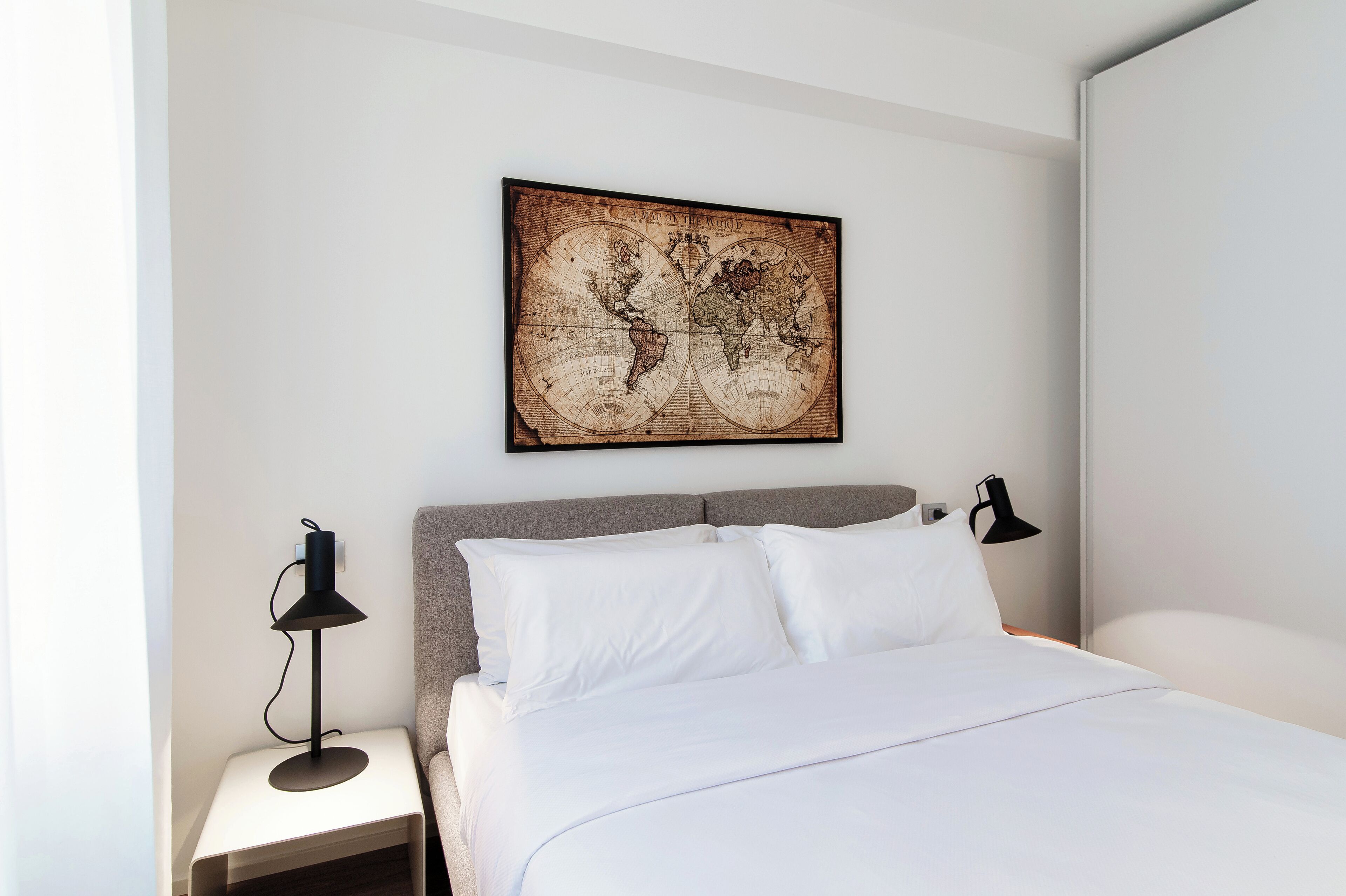 Een net opgemaakt bed met witte lakens, een wereldkaart aan de muur en een bedlamp op een nachtkastje.