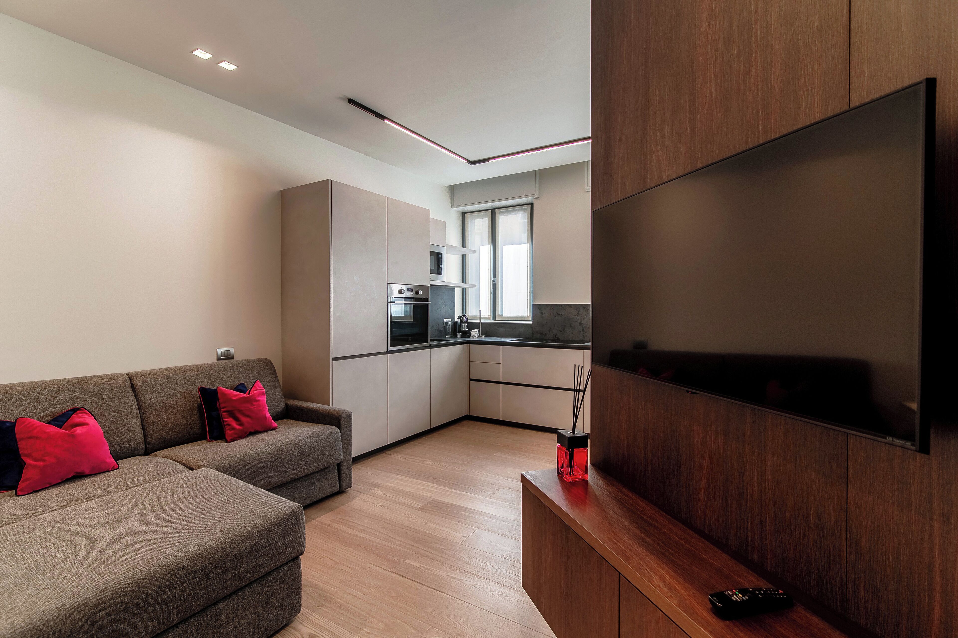 A modern living room with a sofa, a flat-screen TV mounted on the wall, and a kitchen in the background.