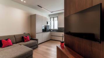 A modern living room with a sofa, a flat-screen TV mounted on the wall, and a kitchen in the background.