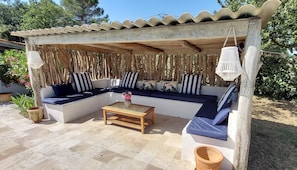 A covered outdoor seating area with a wooden bench, a table, and a potted plant.