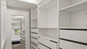 Un dressing accessible sans porte, avec des étagères et des tiroirs blancs, un sol recouvert de moquette et une porte de placard miroir.