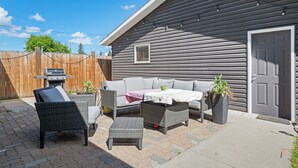 Un espace patio avec une maison grise, des meubles en osier, un barbecue et une plante en pot.