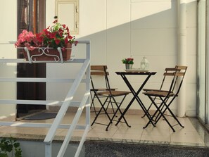 A balcony with a small table and two chairs, a potted plant with flowers, and a view of a building.