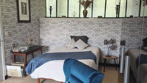Ein Schlafzimmer mit Backsteinwand, einem Bett mit blauer Bettdecke, einem blauen Sessel, einem kleinen Tisch mit Lampe und einem Spiegel.