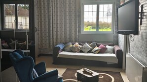 Ein modernes Hotelzimmer mit Schlafsofa, blauem Sessel, flachbildfernseher an der Wand und einem großen Fenster mit Blick ins Freie.