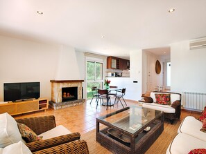 A living room with a television, fireplace, wicker furniture, and a glass coffee table. - Beautiful Villa in Sant Feliu de Guíxols with Shared Pool (Sant Feliu de Guíxols)