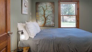 Una camera da letto con un letto, un comodino, una lampada, una finestra con persiane e un quadro raffigurante un albero.