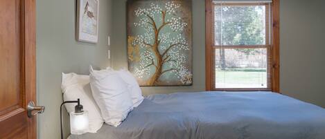 A bedroom with a bed, a nightstand, a lamp, a window with blinds, and a painting of a tree.