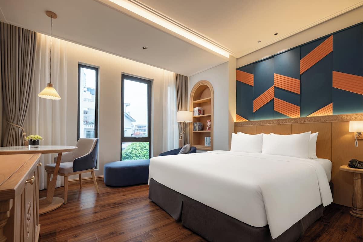 A hotel room with a large bed, a desk, a chair, a bookshelf, and a view of the city through the window.