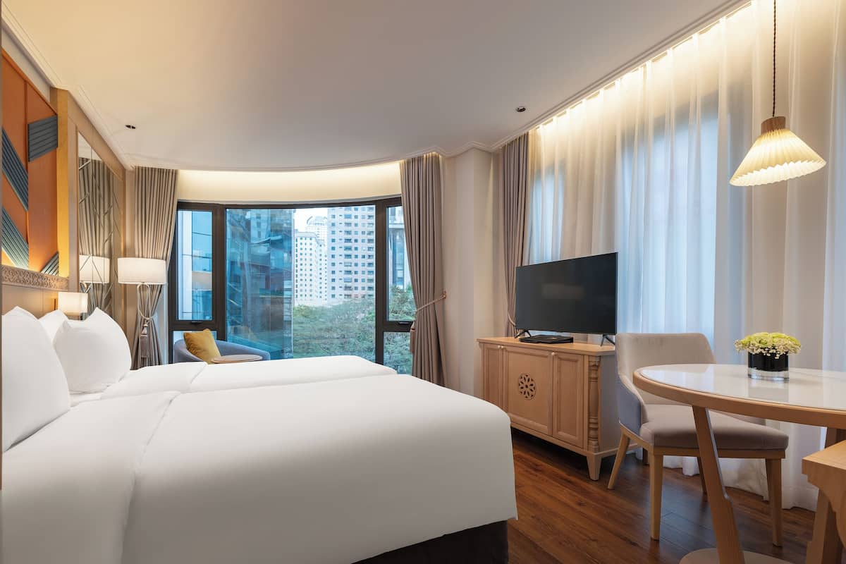 A hotel room with a large bed, a TV on a wooden cabinet, a dining table with a chair, and a view of tall buildings through the window.