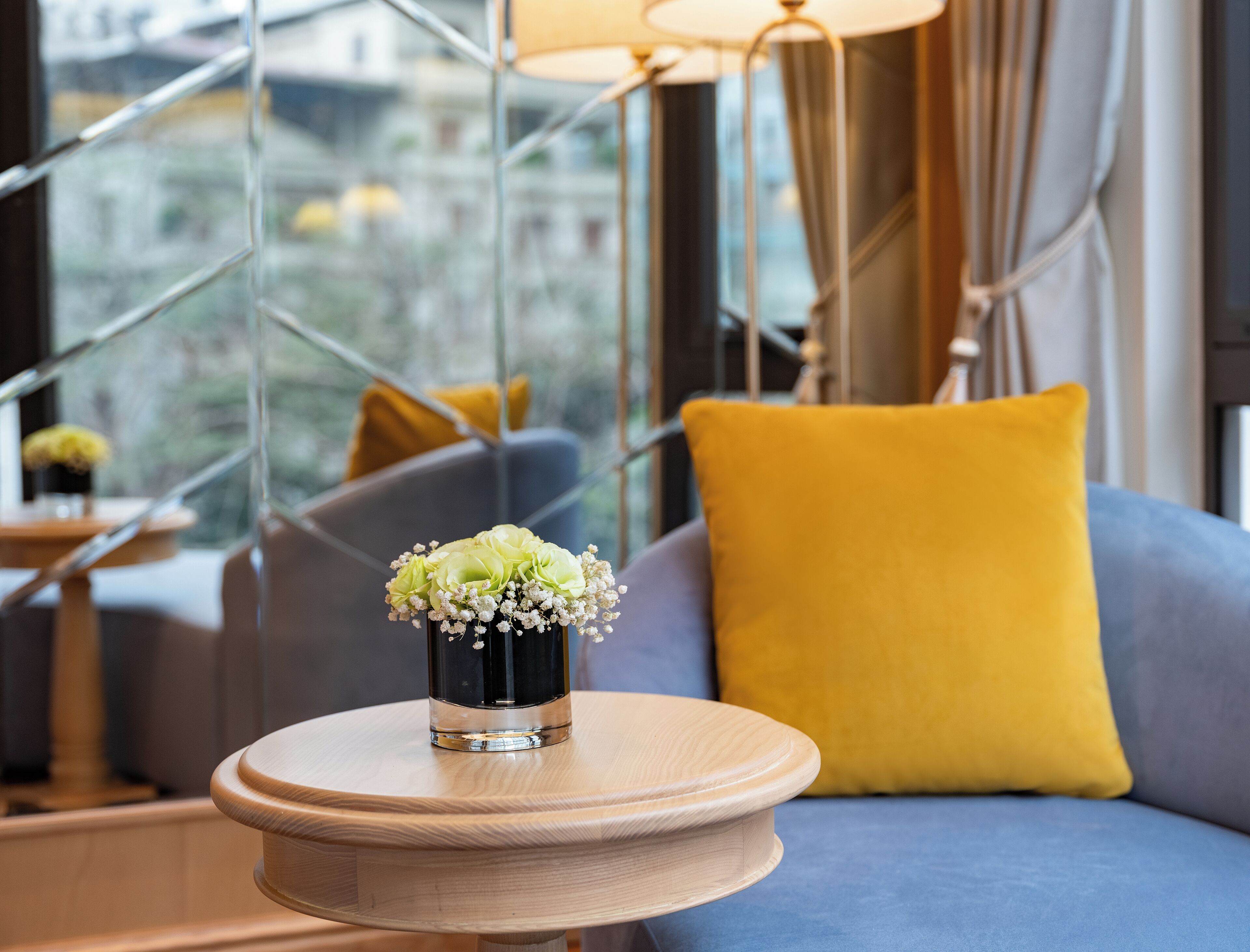 A modern interior with a round wooden table holding a vase with flowers, a blue sofa with a yellow pillow, and a view through large windows.