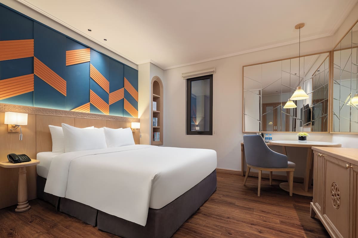 A hotel room with a large bed, a desk, a chair, a phone on the nightstand, and a bookshelf.