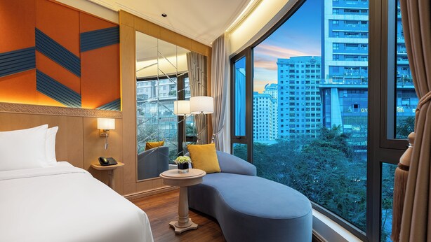 A hotel room with a bed, a seating area, a small table with a lamp, and a large window with a city view. - Saigon Aroma Hotel (Hanoi)