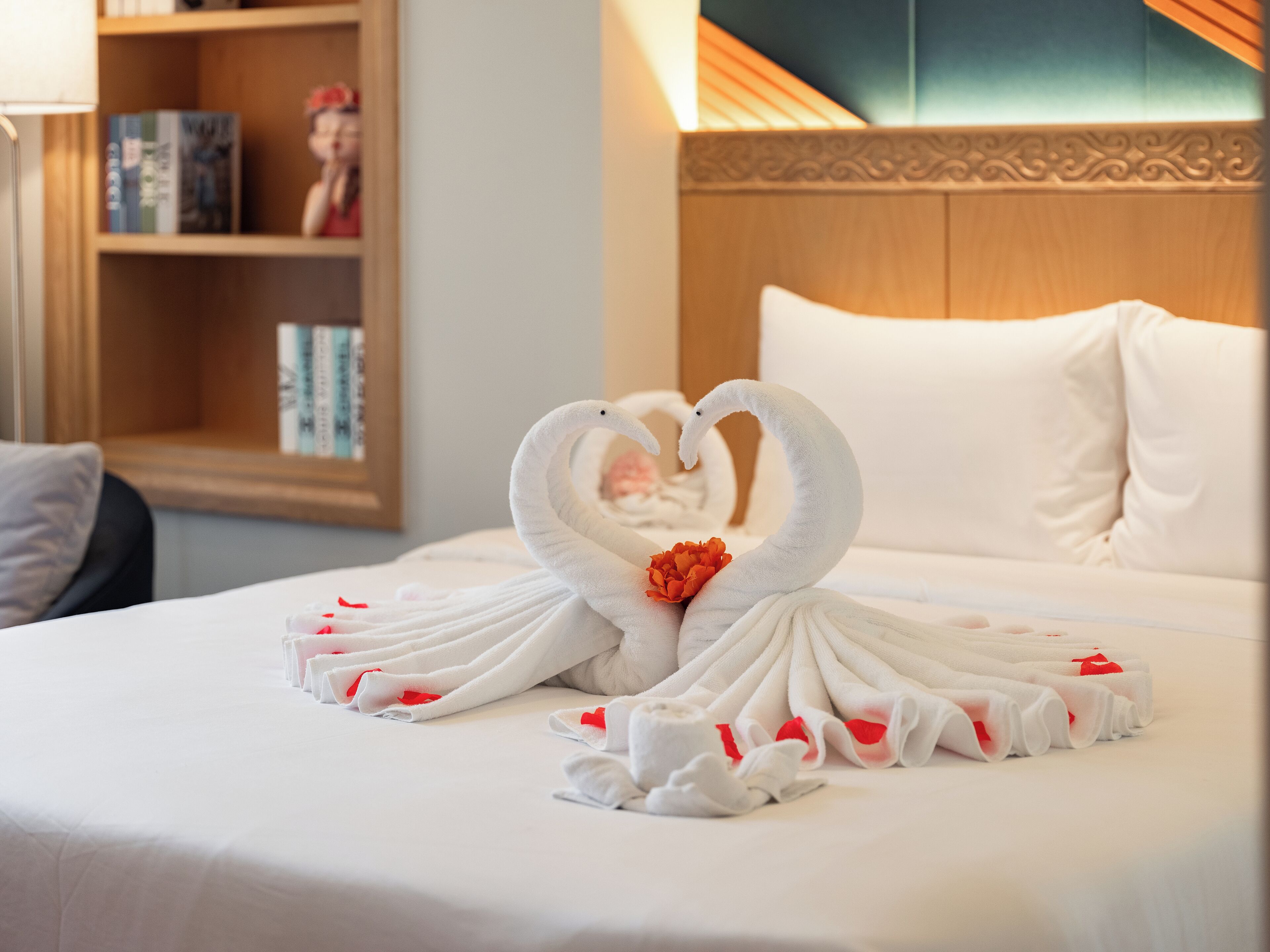 A neatly made bed with a heart-shaped towel arrangement and a single red flower.