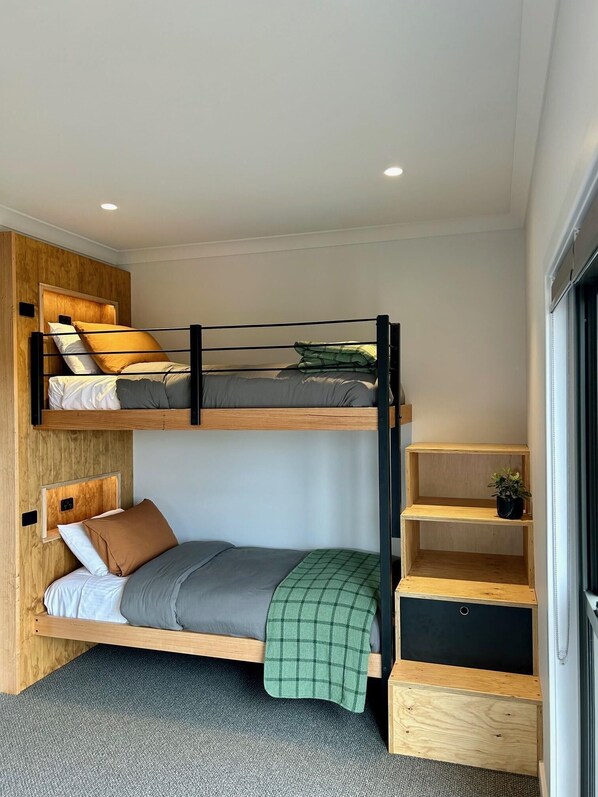 Habitación con literas, cabecera de madera, mesita de noche y una pequeña estantería con una planta.