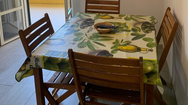 A dining area with a table covered by a tablecloth with a fruit pattern, surrounded by wooden chairs. A television is mounted on the wall above the table.