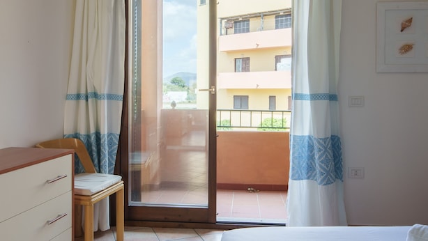 Une chambre à coucher comprenant un lit, une chaise en bois, une commode et une porte-fenêtre coulissante donnant sur un balcon.